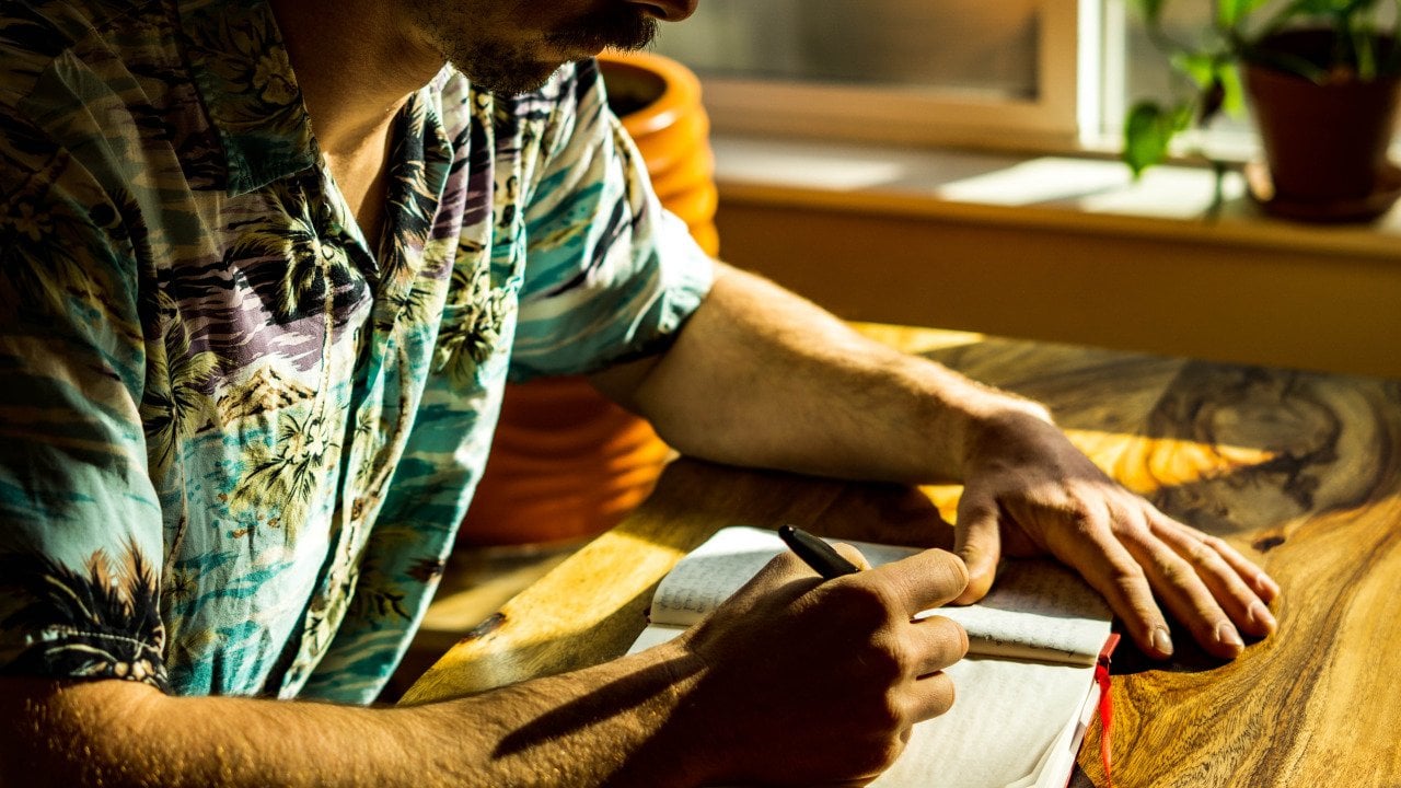 Man wearing a Hawaiian-style shirt sitting next to the window with light coming in, writing in a journal.
