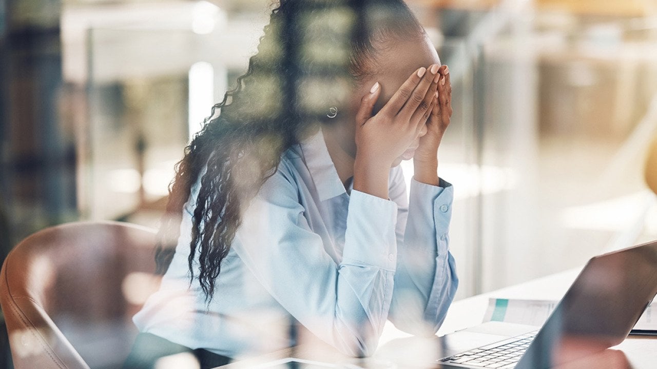 A stressed young black woman with her hands over her face at work.