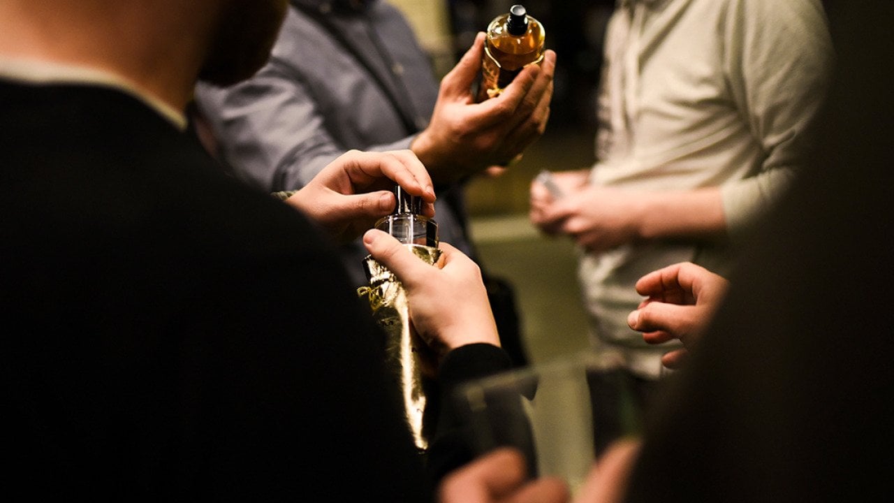 Three men in a room holding perfume bottles and trying out their scents.