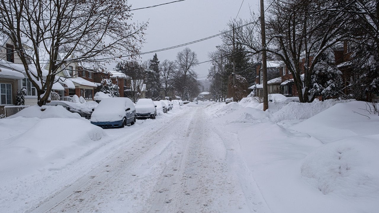 Unplowed snow accumulated on a residential street where rows of cars are parked.
