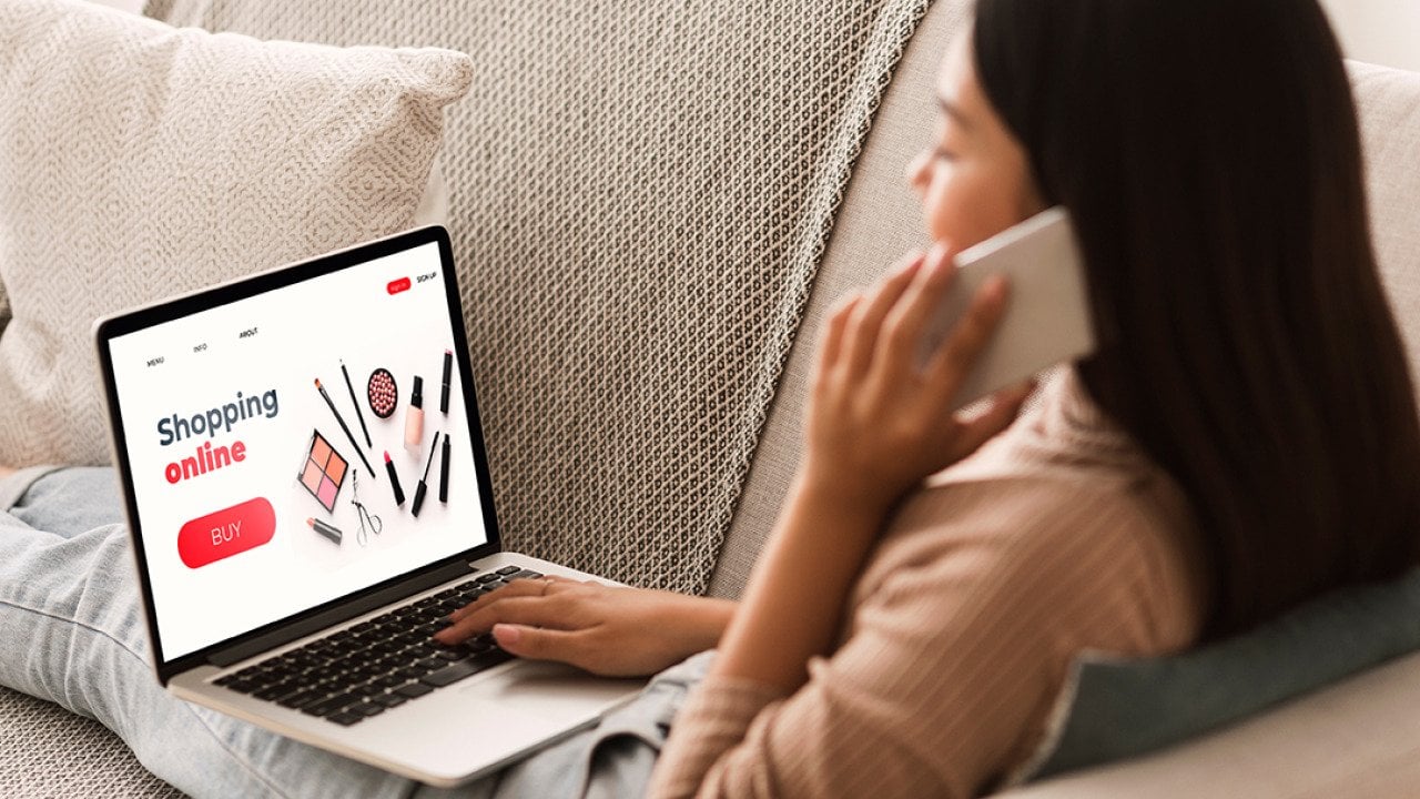 Back view of a woman sitting at home, on the phone, and browsing a shopping website on a laptop.