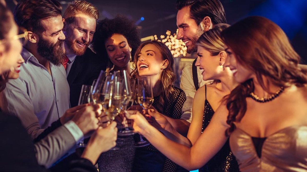 A group of people in party dresses and suits clink glasses together in a party setting.