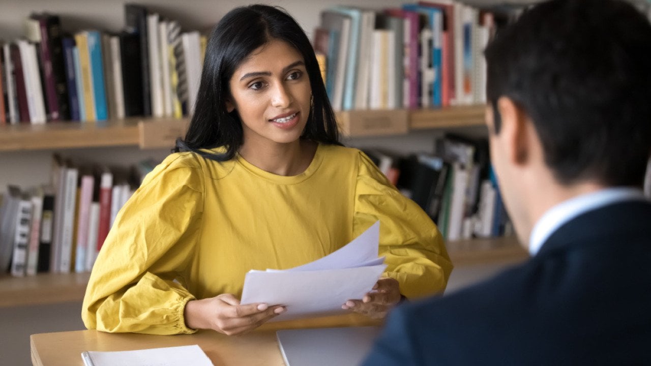 A woman behind a table holding papers and talking to a man sitting on the other side.