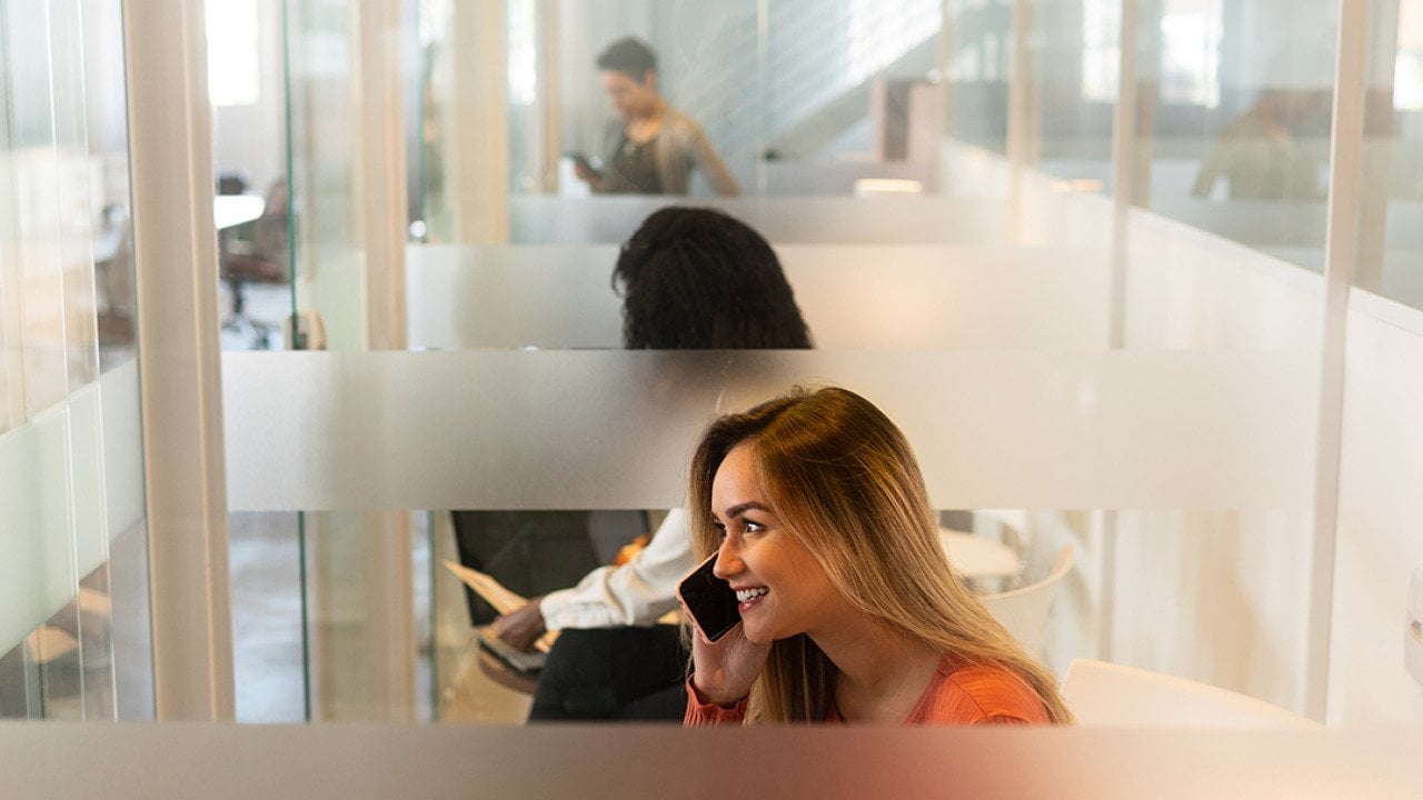 A woman at an office's phone booth taking a call with her smartphone.