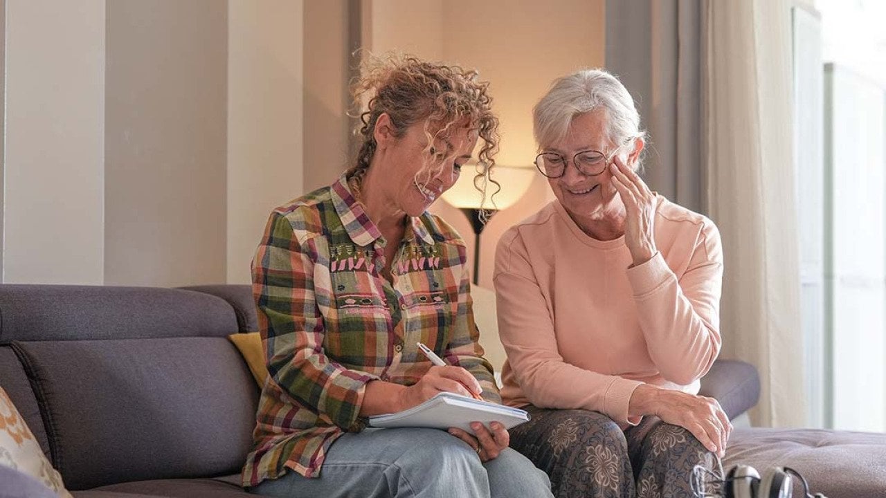 A senior woman and her caregiver sitting together on a sofa at home.