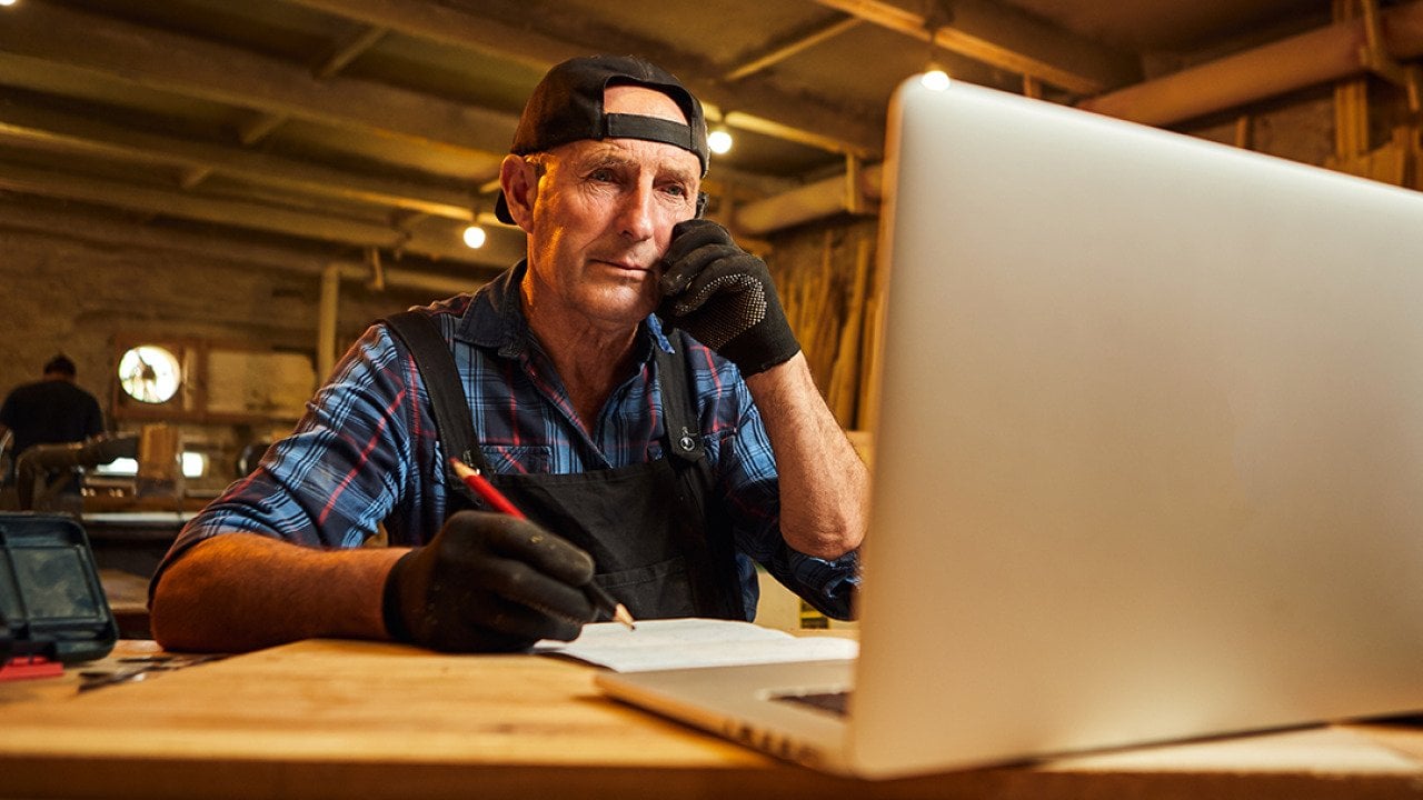 A senior male carpenter working with a client online from their workshop.
