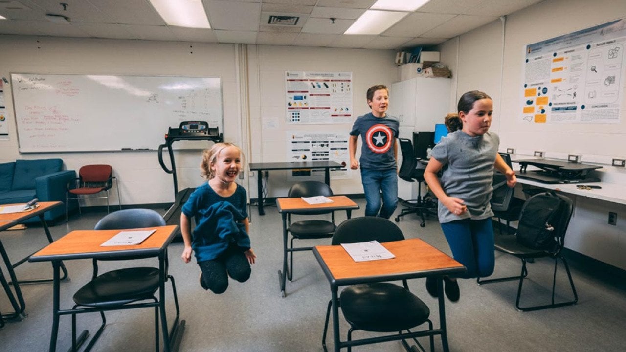 Three elementary-school-age students jumping in the air next to desks in a classroom.