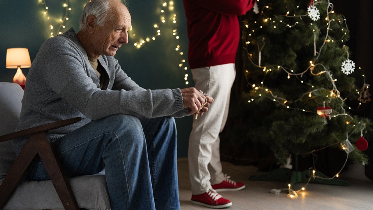 A stressed elderly man sitting by their Christmas tree as his partner decorates it.