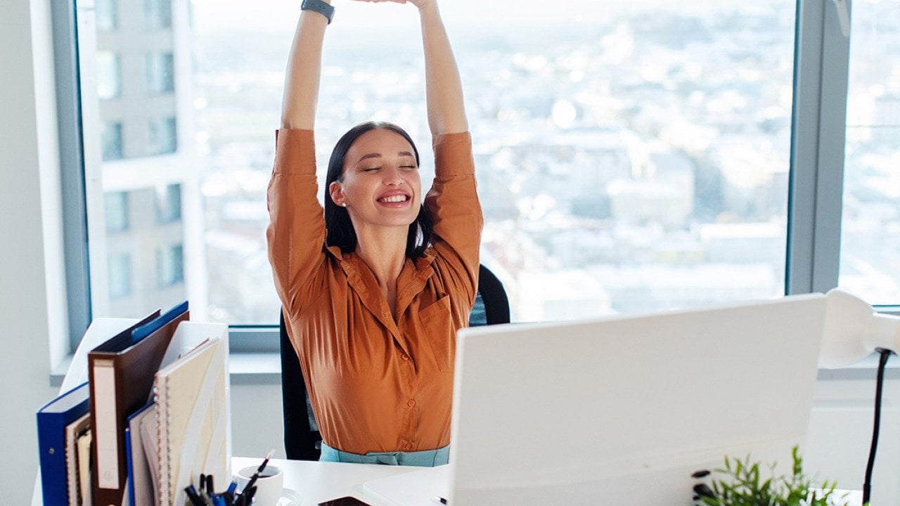 A female manager happily stretching her back and arms while seated at her desk in an office.