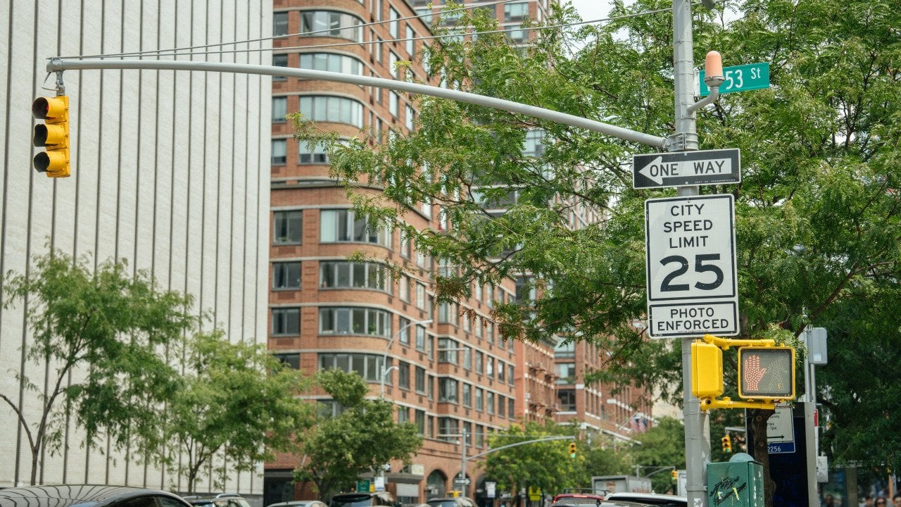 A City Speed Limit 25 sign with a photo enforcement notice stands near a traffic light on a busy urban corner. Modern buildings, cars, and a street sign for 53rd Street are visible in the background.