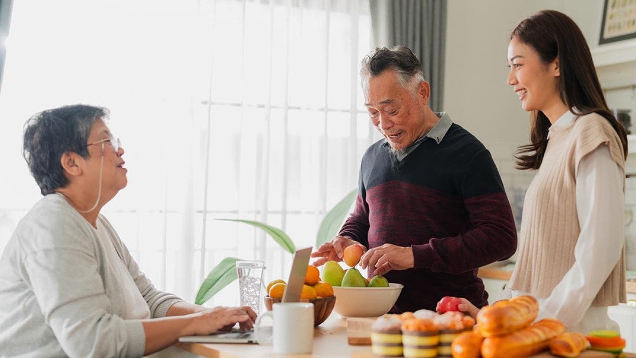 Mother father and adult daughter at the kitchen table talking with window in the background and fruit on the table.