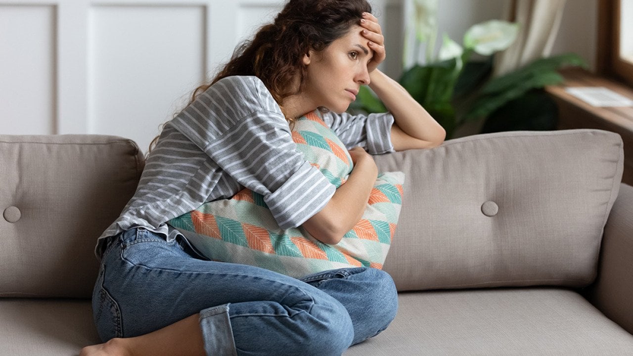A stressed young woman, sitting at a sofa and cuddling a pillow while lost in thought.
