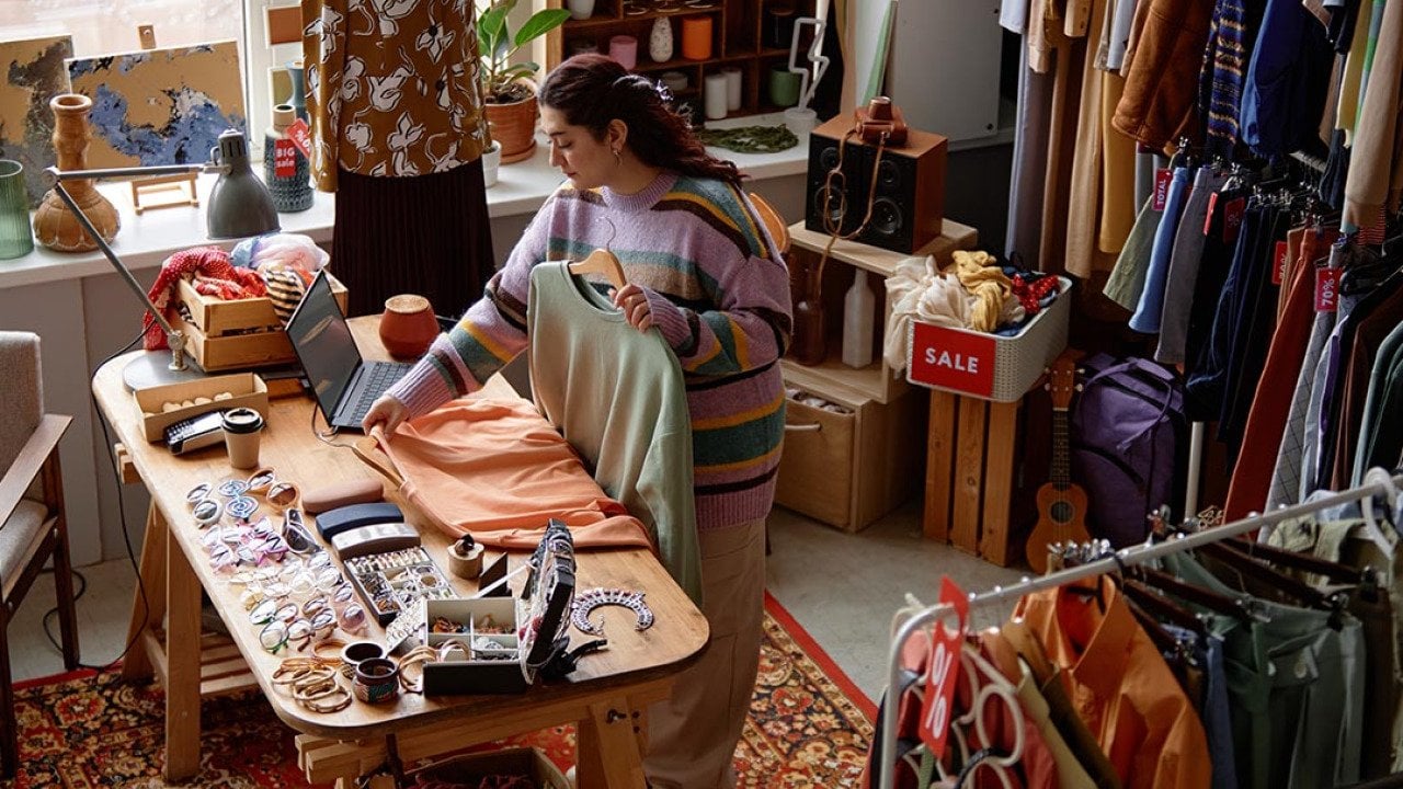 High-angle shot of sales assistant working at checkout desk cluttered with fashion accessories at second hand shop with retro style carpet selling clothes and household goods.