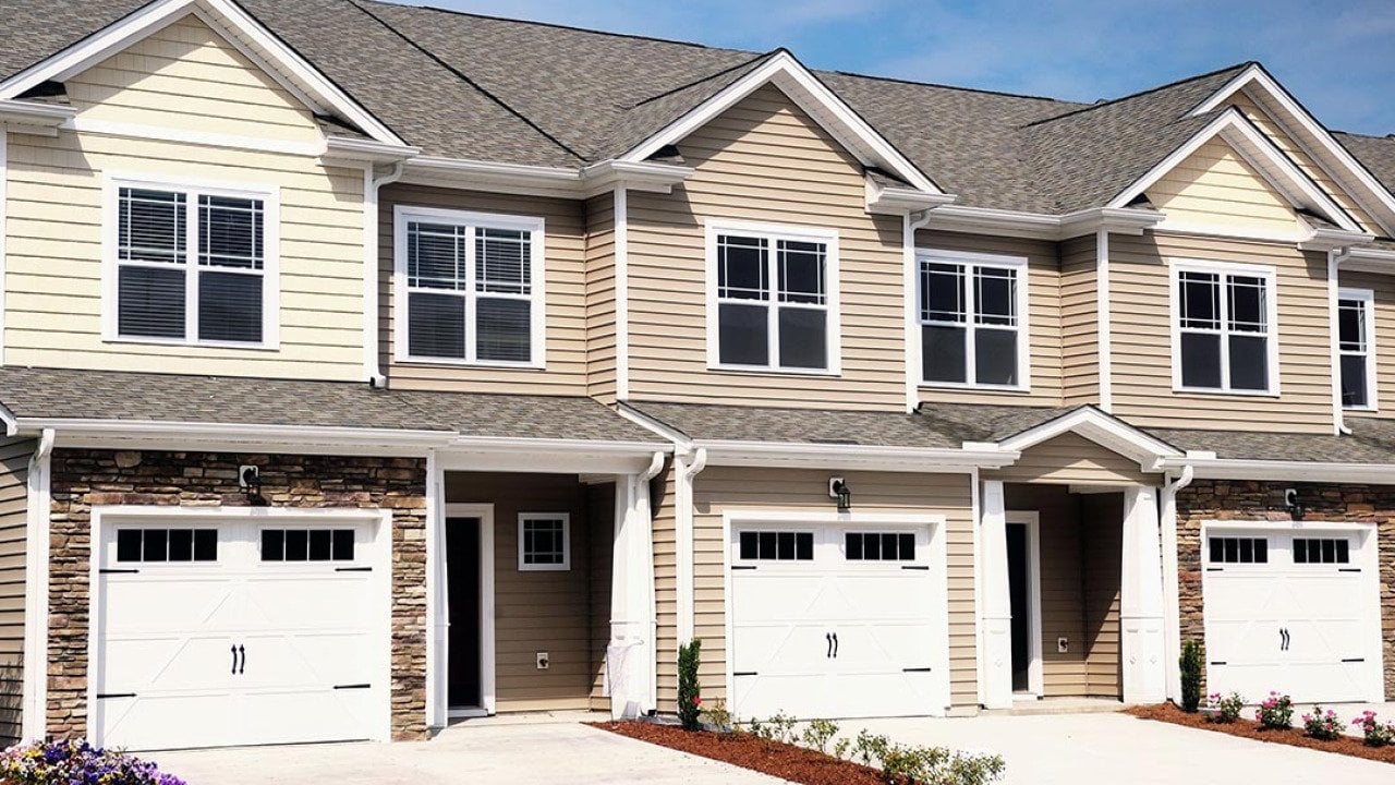 A row of modern townhomes with beige siding, white trim, and stone accents, each featuring a garage, driveway, and small landscaped front yards under a bright blue sky.