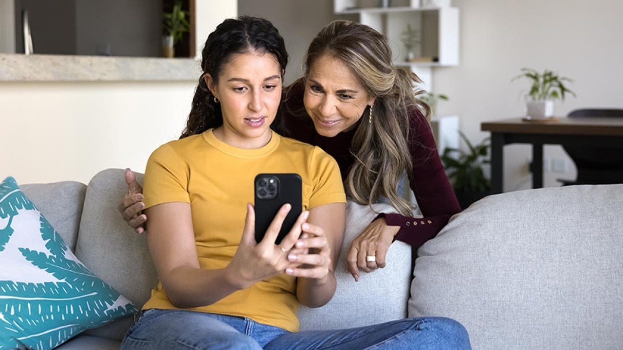 Adolescent daughter on sofa shows interested mum social media on phone screen.