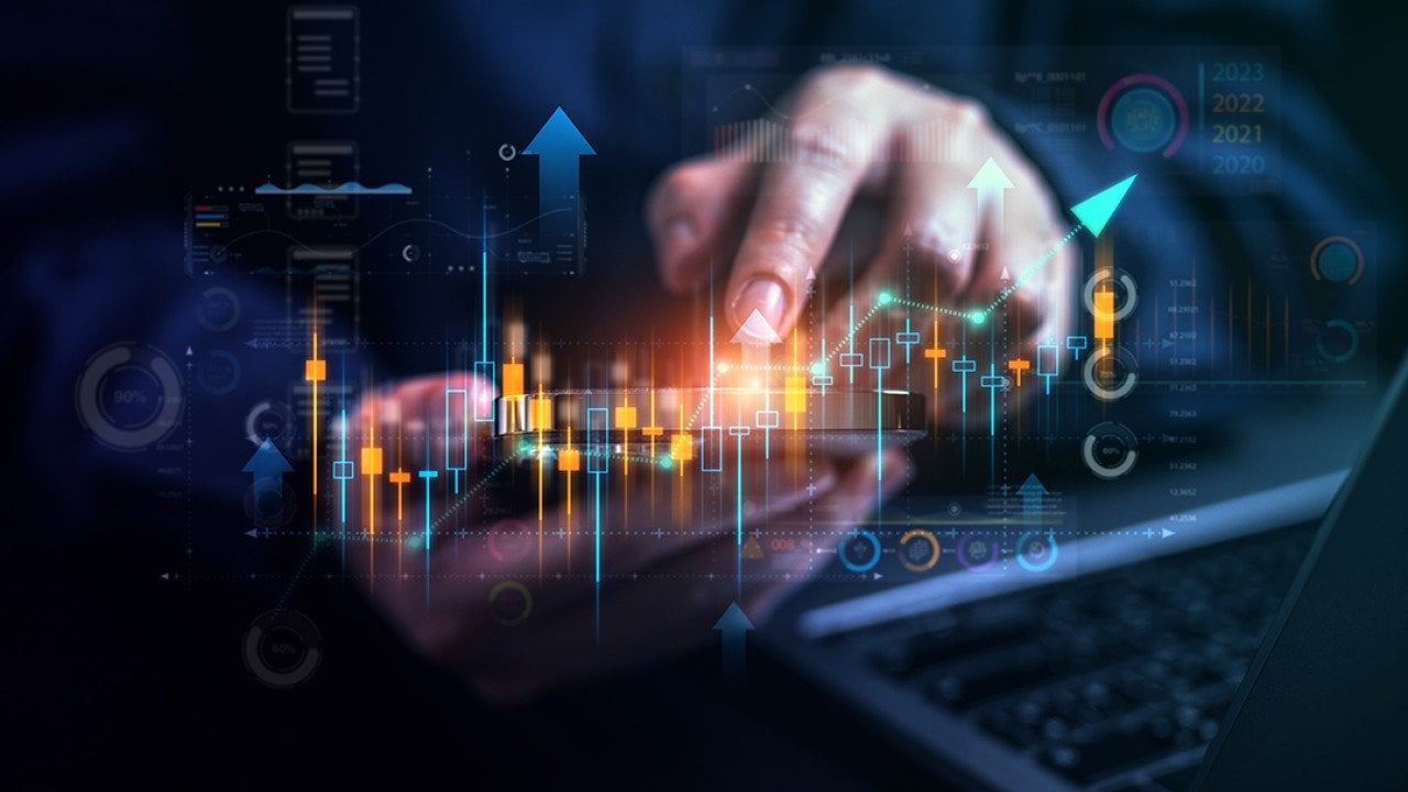 Stock market trading graphics illustrated over a businessperson using a phone and laptop.