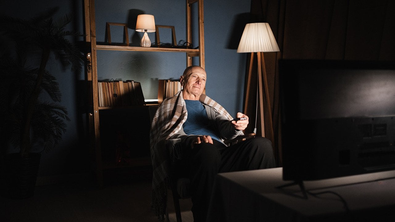 An elderly man sitting in a dim living room and watching TV.
