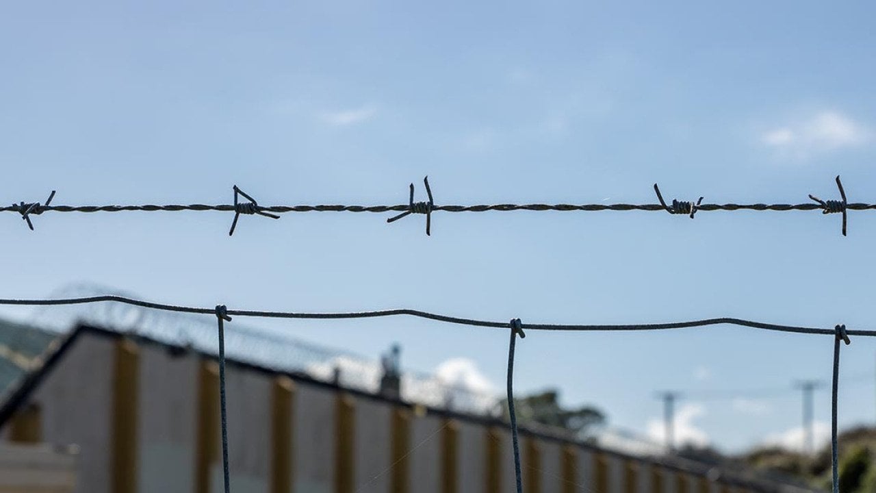 Close up of a single strand of barbed wire above a fence with a correctional facility in the background.