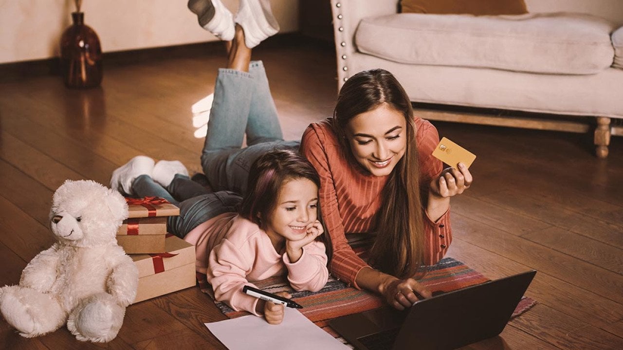 Girl writing wishlist, next to adult woman, both are laying on a rug looking a laptop online shopping while the woman is holding a credit card. There are wrapped gifts next to them along with a teddy bear.