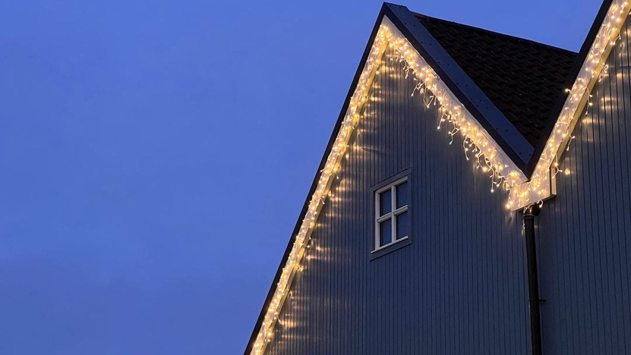 Icicle Christmas Lights hanging along the roof line of a wooden-fronted house. There is a small white window on the house, and a dusky blue sky behind the house.