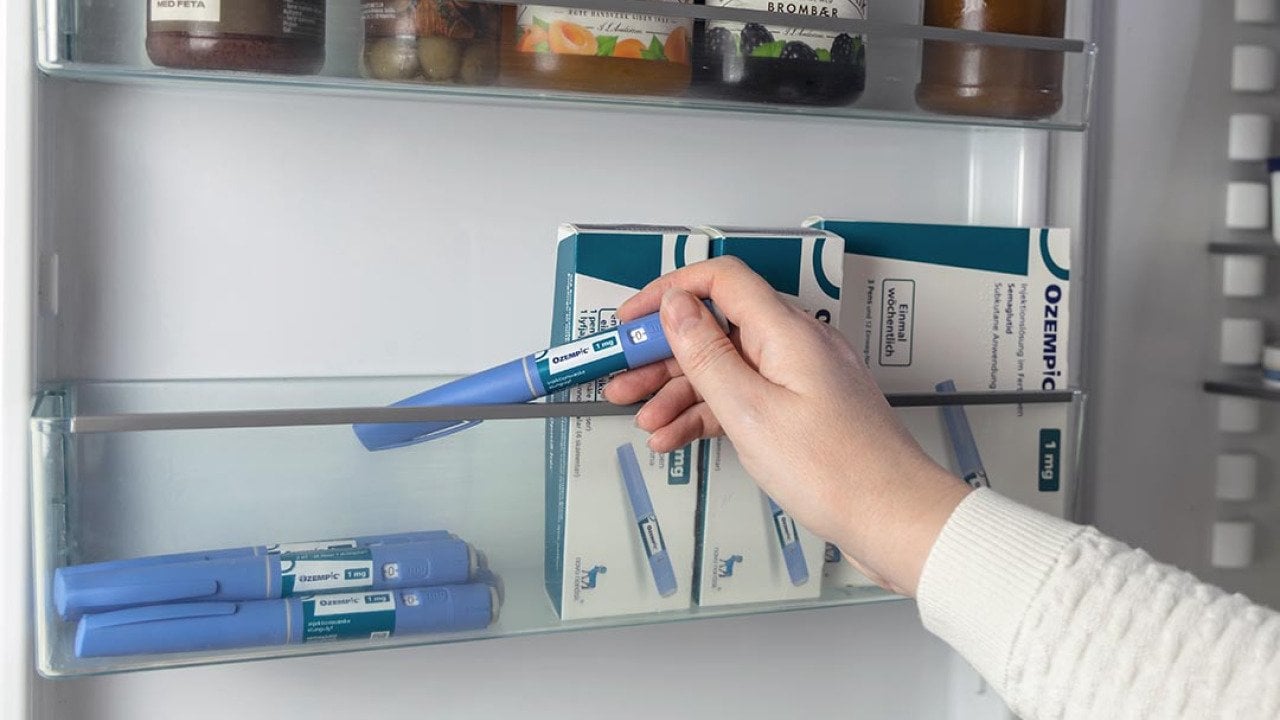 Close up of arm of person wearing white sweater reaching into refrigerator door to pick up an Ozempic injection pen, boxes of Ozempic are stored to the right.