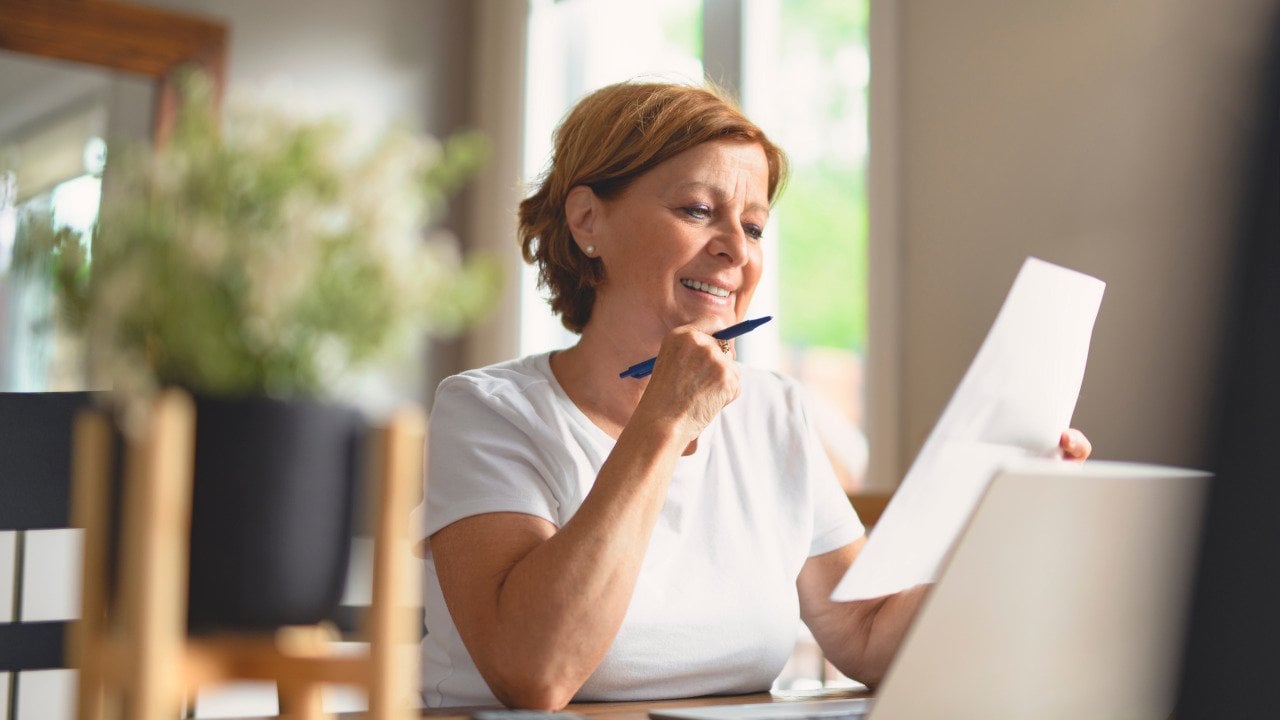 Middle-aged woman sitting at a table looking over paperwork with laptop open and pen in hand.