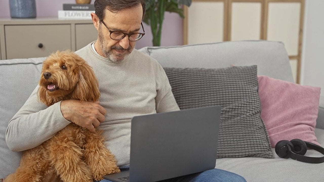 A hispanic middle-aged man cuddles a poodle while using a laptop in a cozy living room setting.