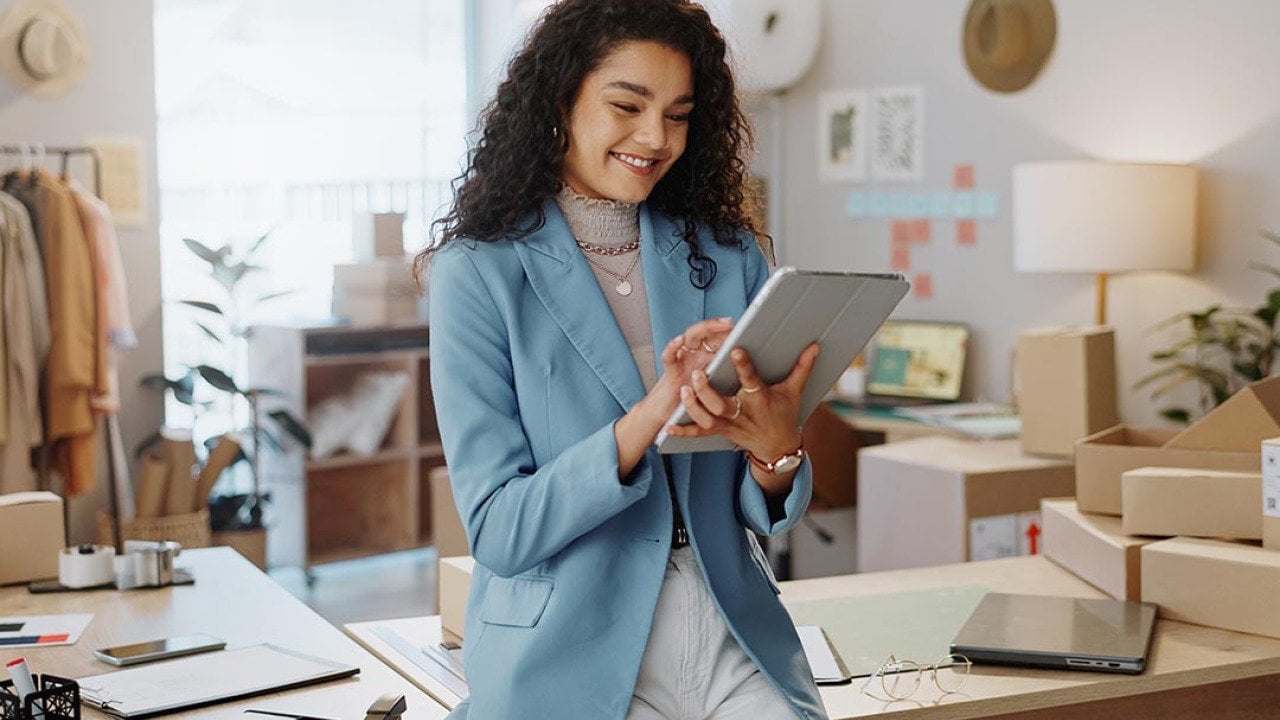 Fashion designer wearing powder-blue oversized blazer and white pants uses a tablet and tablet working logistics or small business managementwith office, shipping and clothing rack behind her.