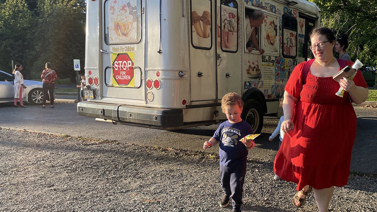 Kimberly Halevy wearing a crimson red midi dress with mid-length sleeves walks with her son Joshua beside her, enjoy treats from an ice cream truck visible behind them, after a recent session with a special instructor.