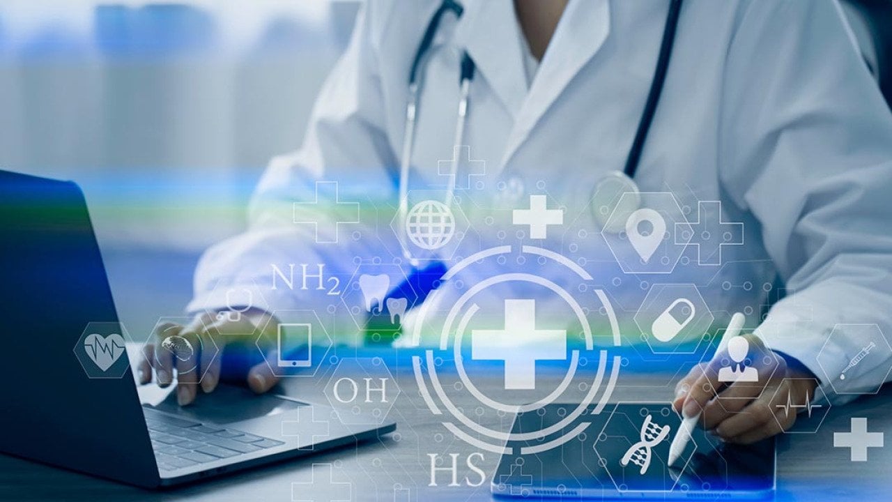 Doctor visible from the neck down sitting at desk with laptop and iPad working with digital medical interface icons superimposed symbolizing AI.