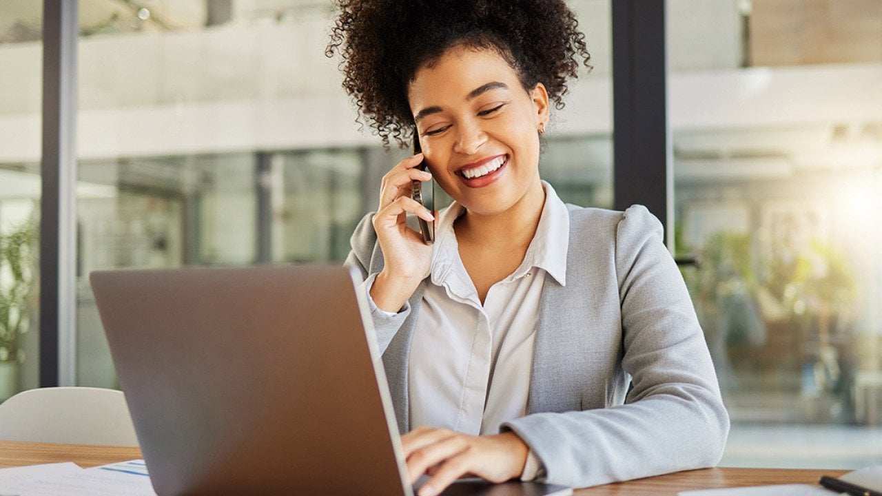 A businesswoman in conversation with someone on her phone and working with a laptop in an office.