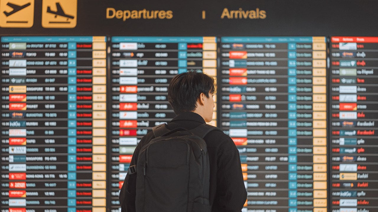 Rear view of a young man with a backpack looking at an airport's flight schedule board.