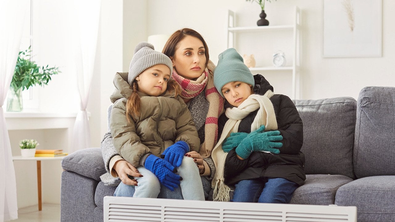 A mother with her two children in winter clothing sitting on a sofa in a cold living room.