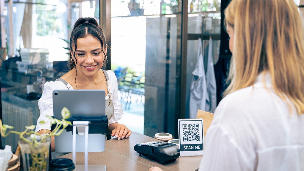 Barista receives a customer lining up to pay in a cafe's counter where a QR code information graphic is displayed.