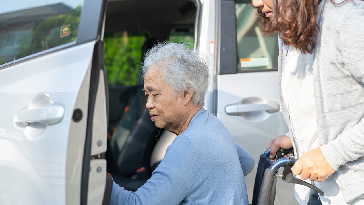 An elderly senior woman in a wheelchair being assisted by her caregiver getting in a car.