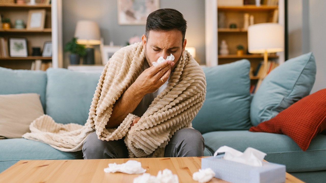 A sick man at home wrapped in blanket sitting on a sofa and blowing his nose.