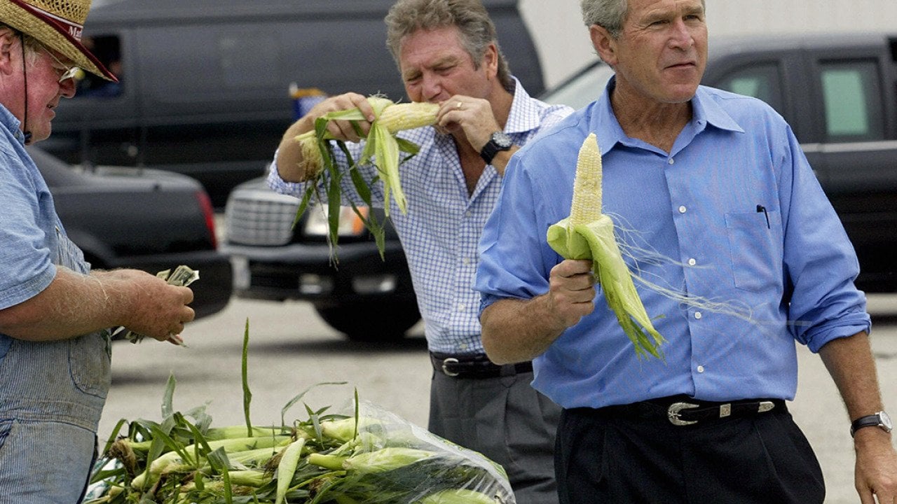 Former US President George W. Bush (R) holding an ear of corn, joined by his friend, Larry Gaitlin (C) while farmer Ken Thompson (L) counts the money handed to him by Bush for a bag of corn, during an impromptu stop of his motorcade at a local farmers market after a campaign rally August 4, 2004 in Davenport, Iowa.