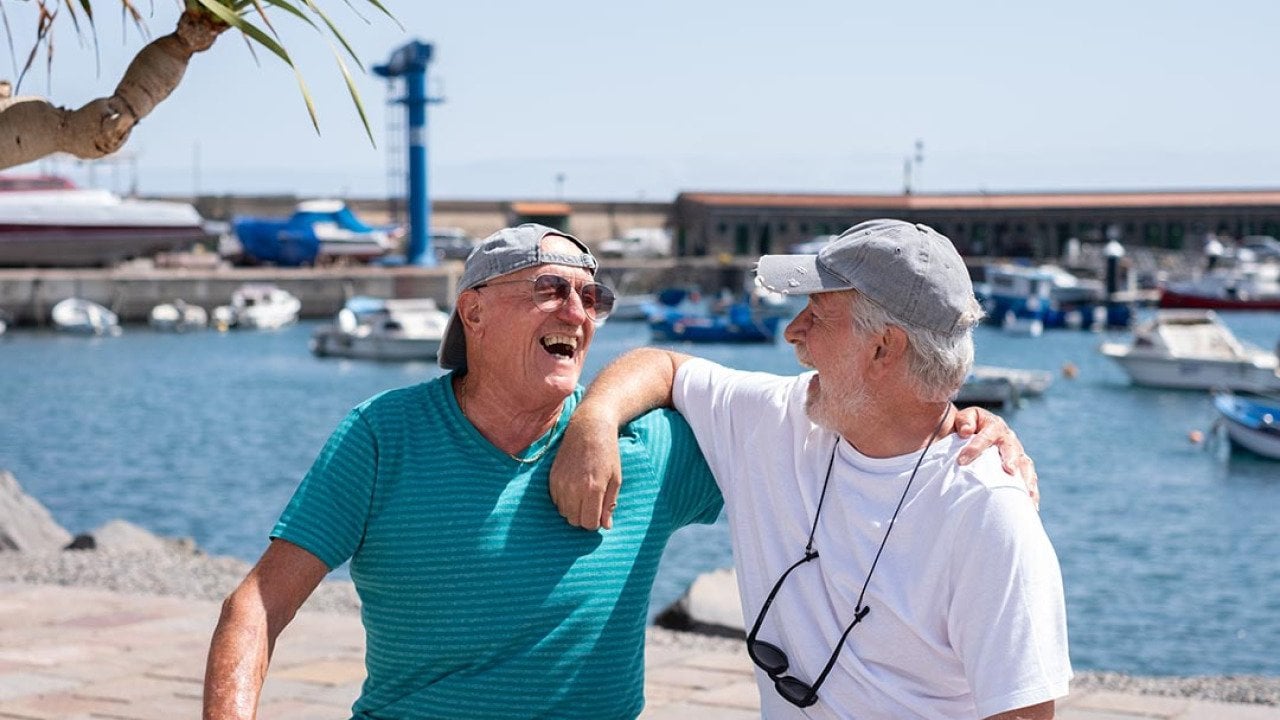 Two senior friends wearing caps and t-shirts in retirement life sitting on a bench at the harbor looking each other laughing happily.