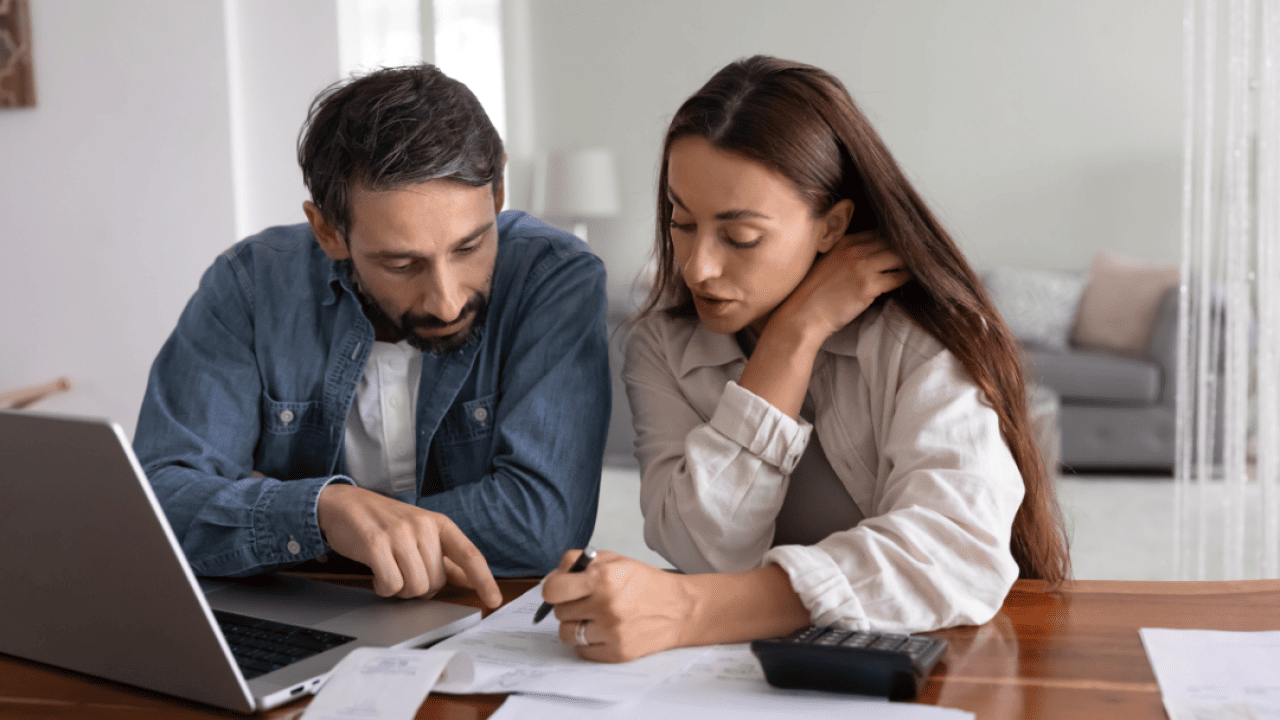 Serious young to middle aged couple discussing expenses at table with papers, a laptop and a calculator.