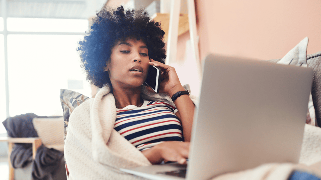 A woman at home on the couch, wrapped in a blanket talking on the phone with a laptop on her lap.