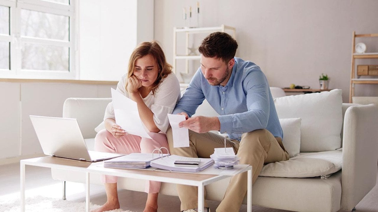 Two people sitting on a white sofa looking at bills and paperwork in a minimalistic living room.