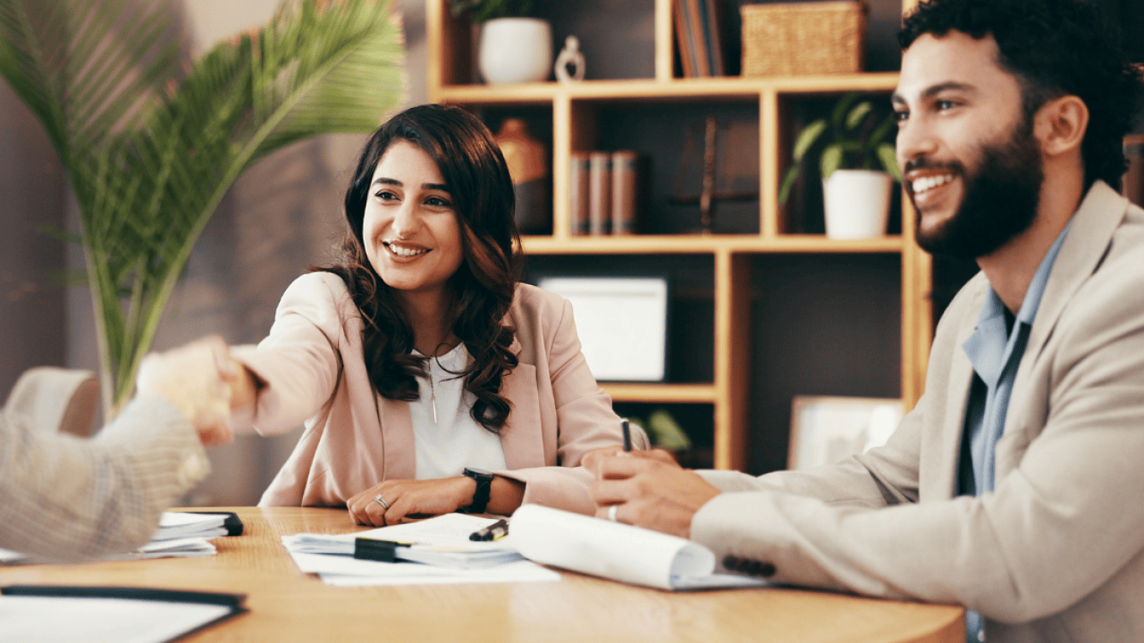 Three business people sit at a table. One smiling person shakes hands with another person, who is mostly out of view. 