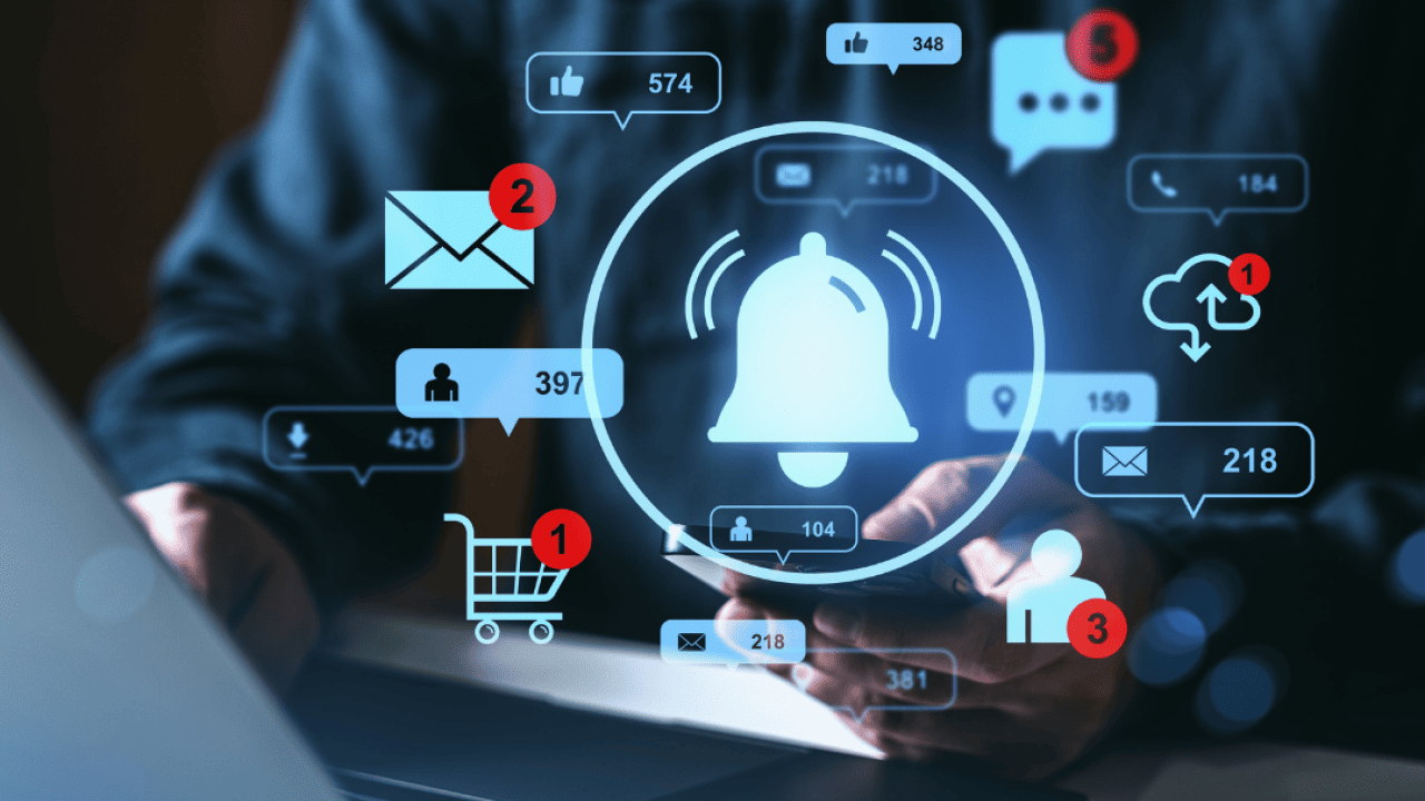 Person using a smartphone in front of a laptop with a various icons, like an alert bell, an email inbox notification, and a shopping cart, overlaying the image. 