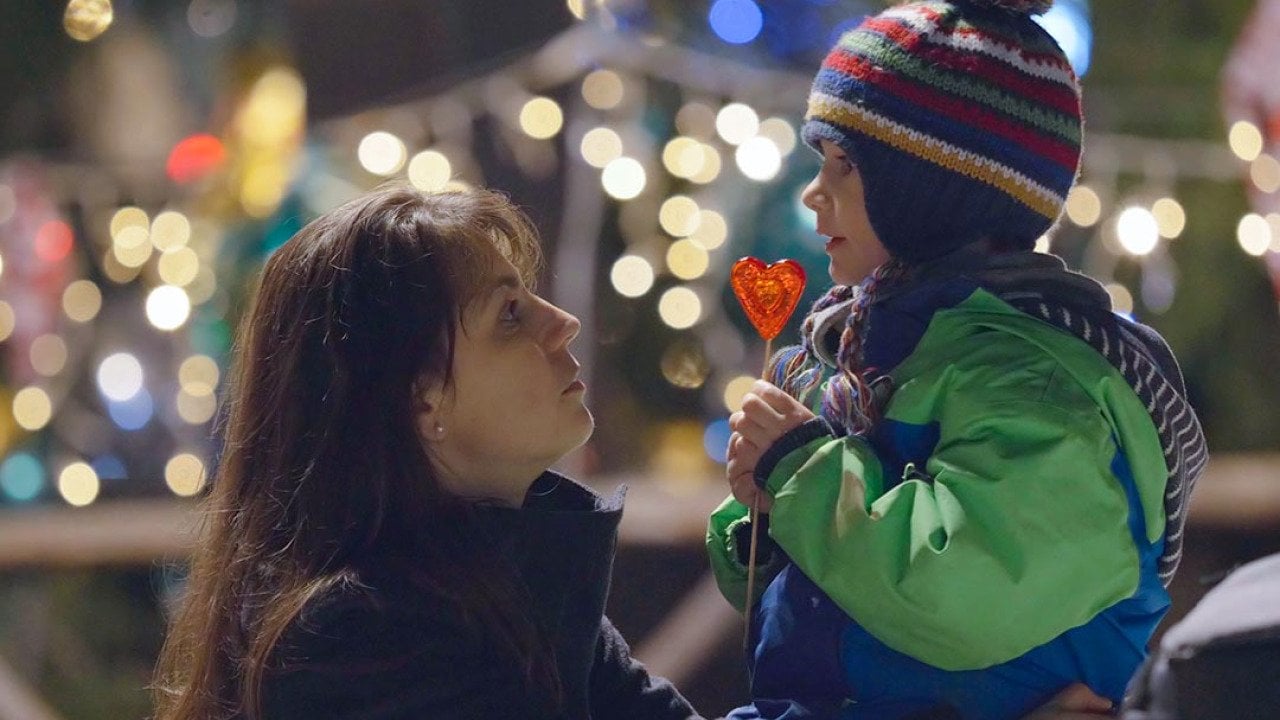 A mother kneels to the height of her child ourdoors and looks into his eyes, he is eating a heart-shaped lollipop and wearing a knitted cap with a pom pom in front of blurry christmas lights in the background.