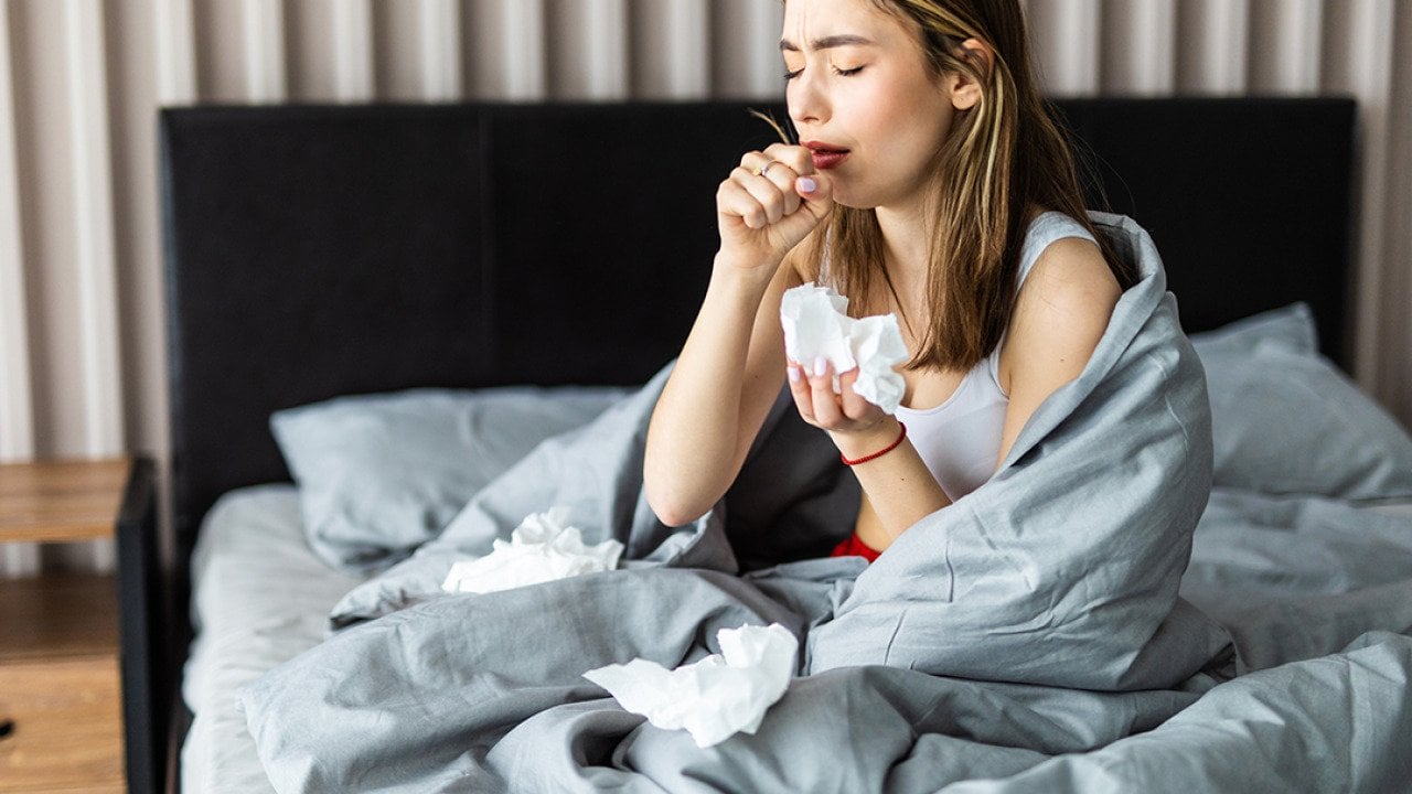 A sick woman wrapped in blanket is sitting on her bed and coughing.