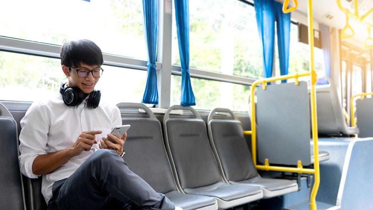 A college-age person wearing a crisp white shirt and headphones around his neck and bright white shoes sits on the side-facing seats in public transportation looking attentively at phone.