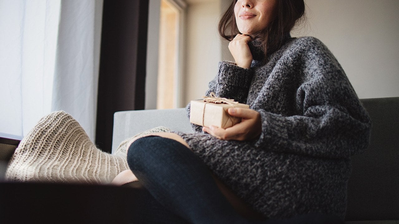 A young woman cozily sitting on a sofa at home holding a small gift box.