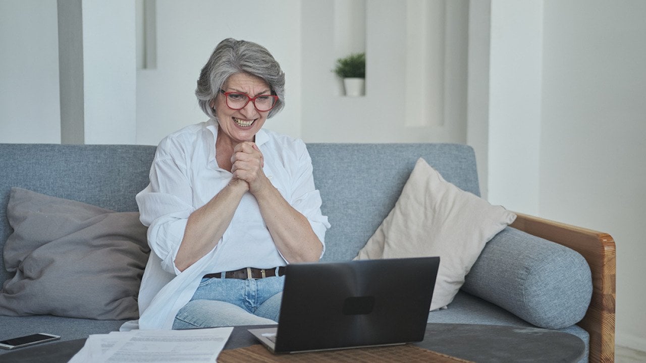 A senior woman sitting on a sofa and excitingly awaits good news on her laptop.