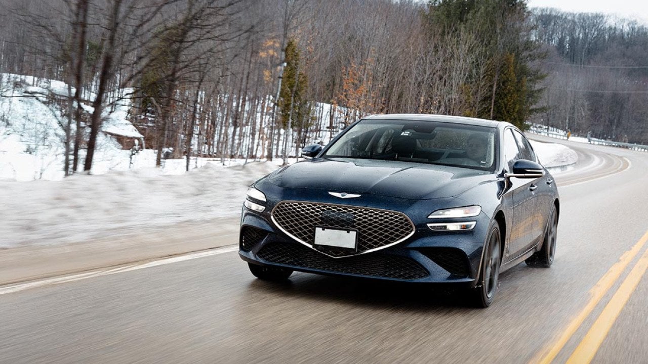 Front view of black Genesis G70 driving on a road in winter with snow and trees in background. 