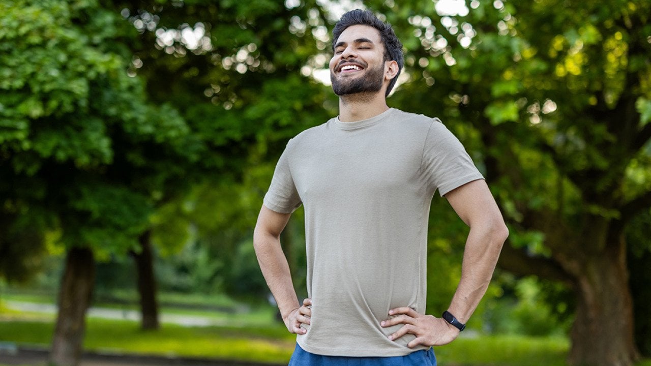 A sporty young man standing outdoors with hands on hips, smiling after an exercise.